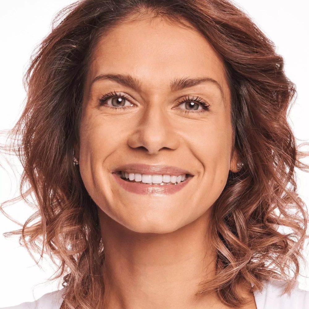 Gros plan d'une femme souriante. Elle a des cheveux bruns ondulés et porte du maquillage. Dents clairement visibles.