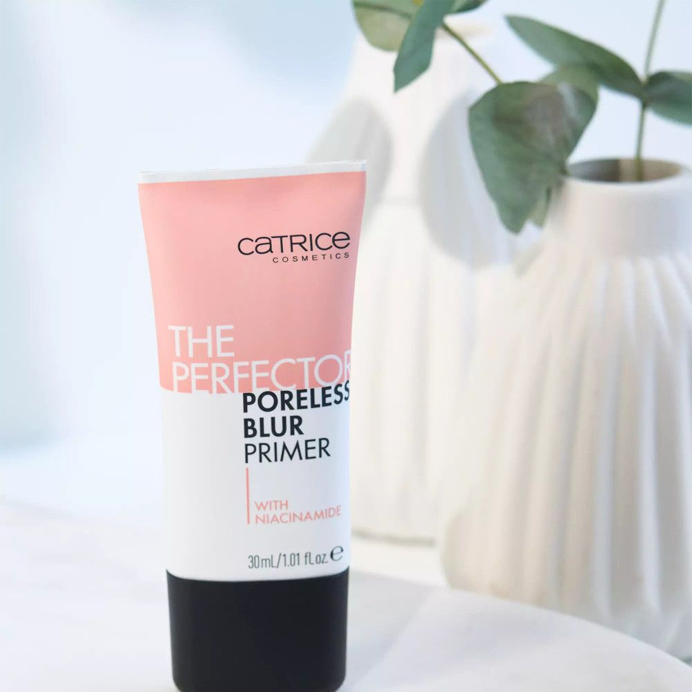 Tube rose et noir. Inscription: Catrice, THE PERFECTOR PORELESS BLUR PRIMER, avec Niacinamide. 30ml/1.01 fl oz e. Devant des vases.