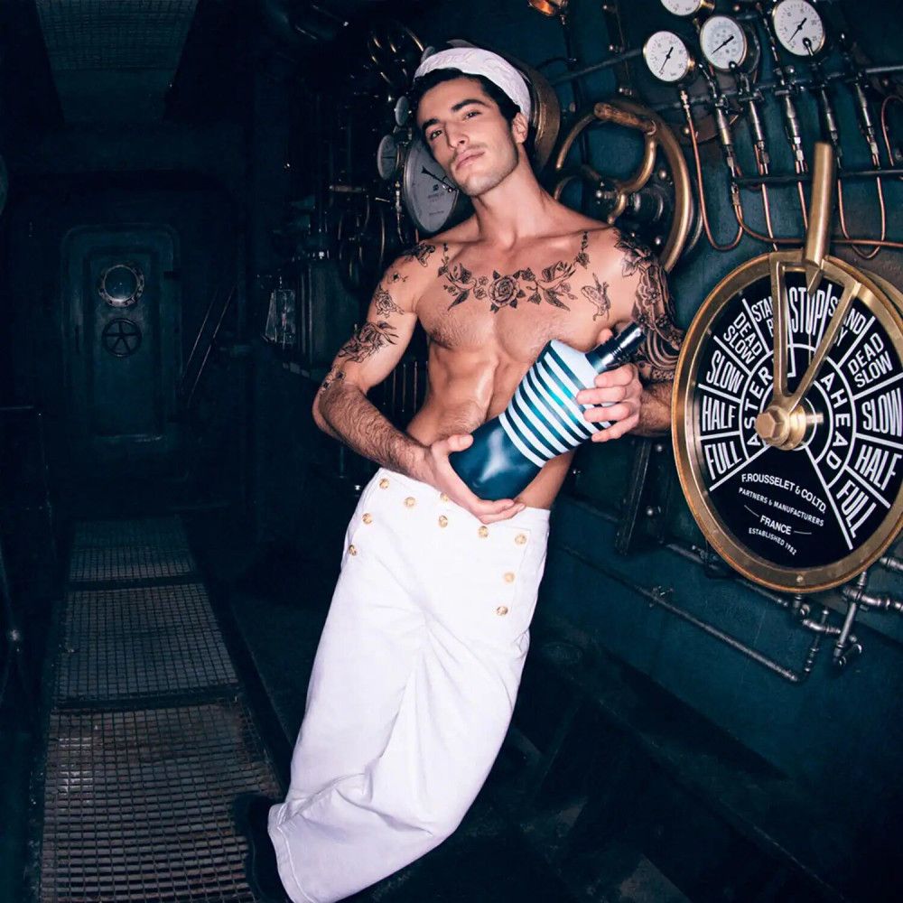 Homme en uniforme de marin tenant un flacon en forme de torse. Flacon rayé bleu et blanc. Fond : salle des machines.