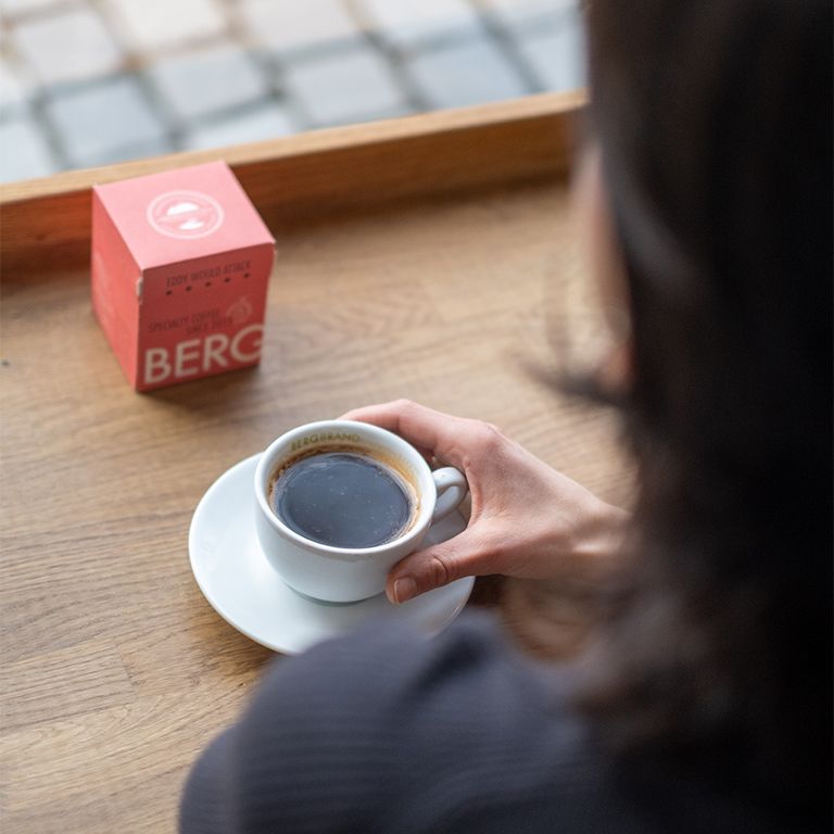 Personne tenant une tasse de café. Sur la table, un emballage rouge. Inscription : BERGBRAND, Kapselkaffee.