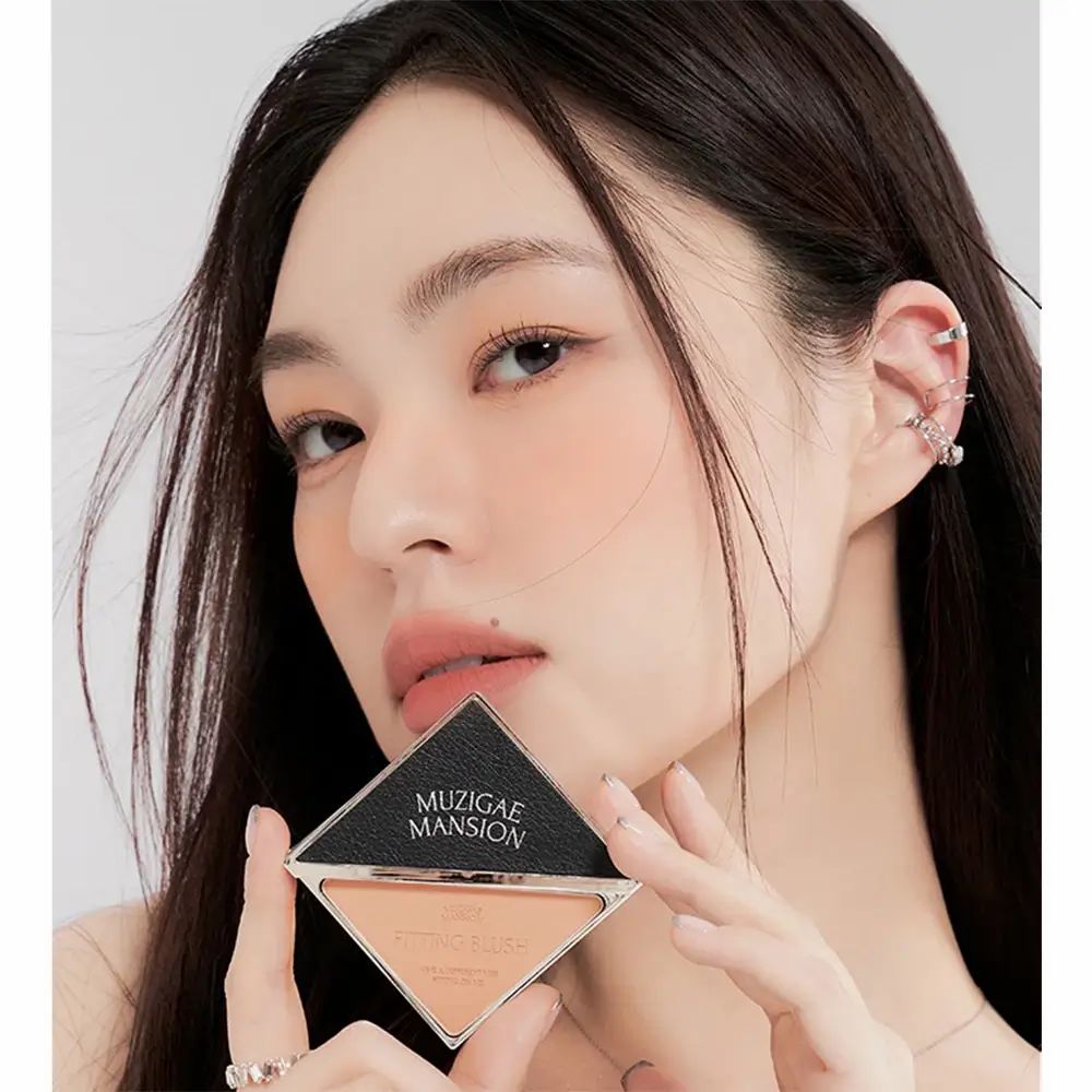 Vrouw houdt MUZIGAE MANSION Fitting Blush vast. Zwarte, driehoekige compacte verpakking. Beige blush. Vrouw met lang, donker haar.