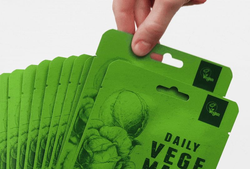 Meerdere groene gezichtsmaskers met opdruk "DAILY VEGE MASK". Een hand grijpt naar een masker. Veganistisch logo.