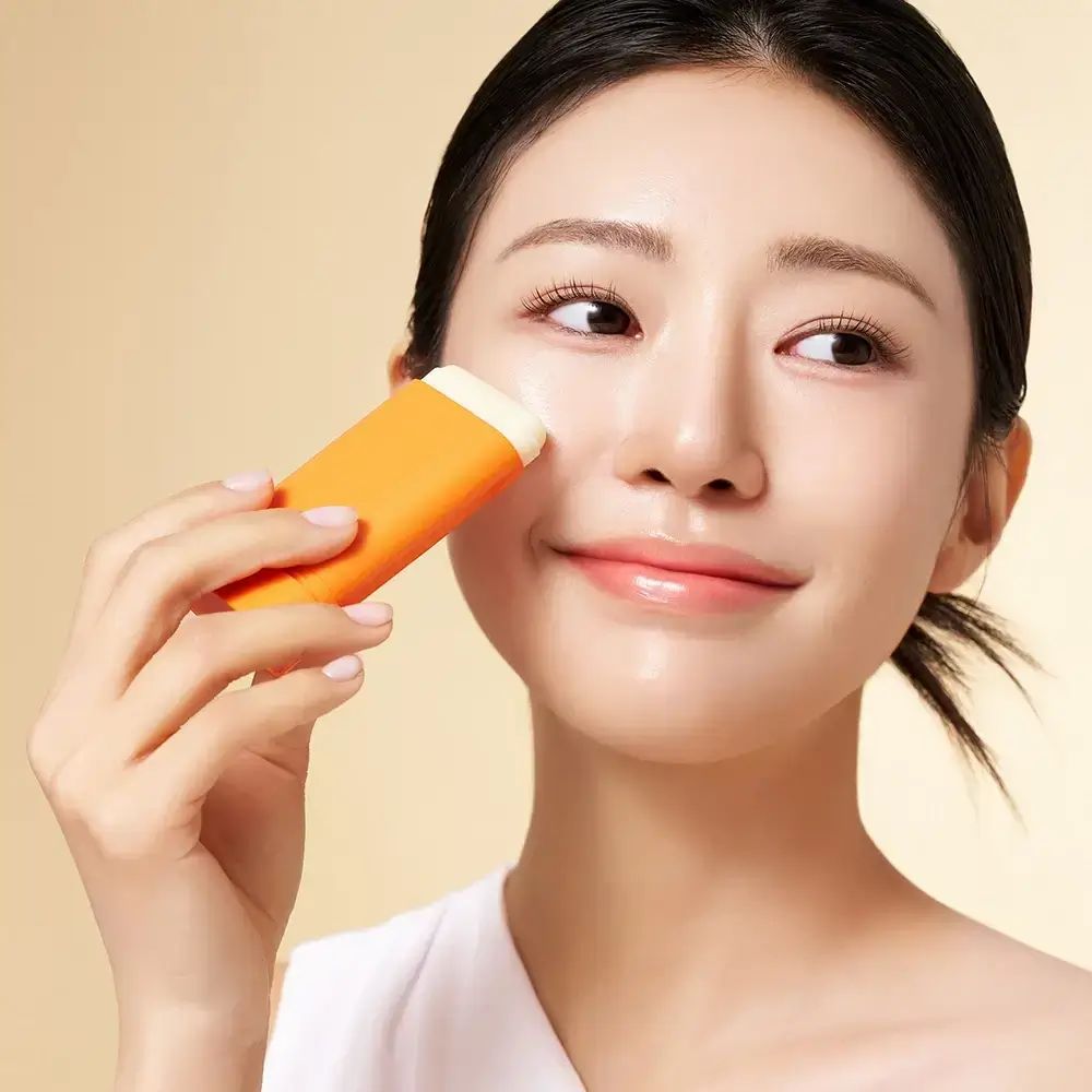Femme appliquant un stick de protection solaire orange sur sa joue. Le stick a une tête blanche. Fond clair.