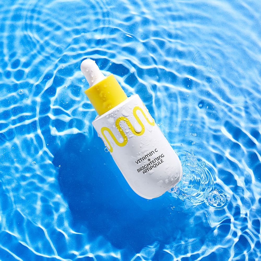 Flacon compte-gouttes blanc avec bouchon jaune, entouré d'eau. Contient Vitamin C Brightening Ampoule.