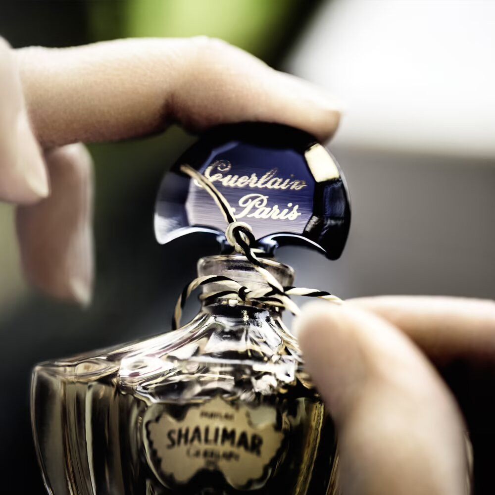 Close-up van een Guerlain Shalimar fles. Handen houden de dop vast. Opschrift: Guerlain Paris, Shalimar.