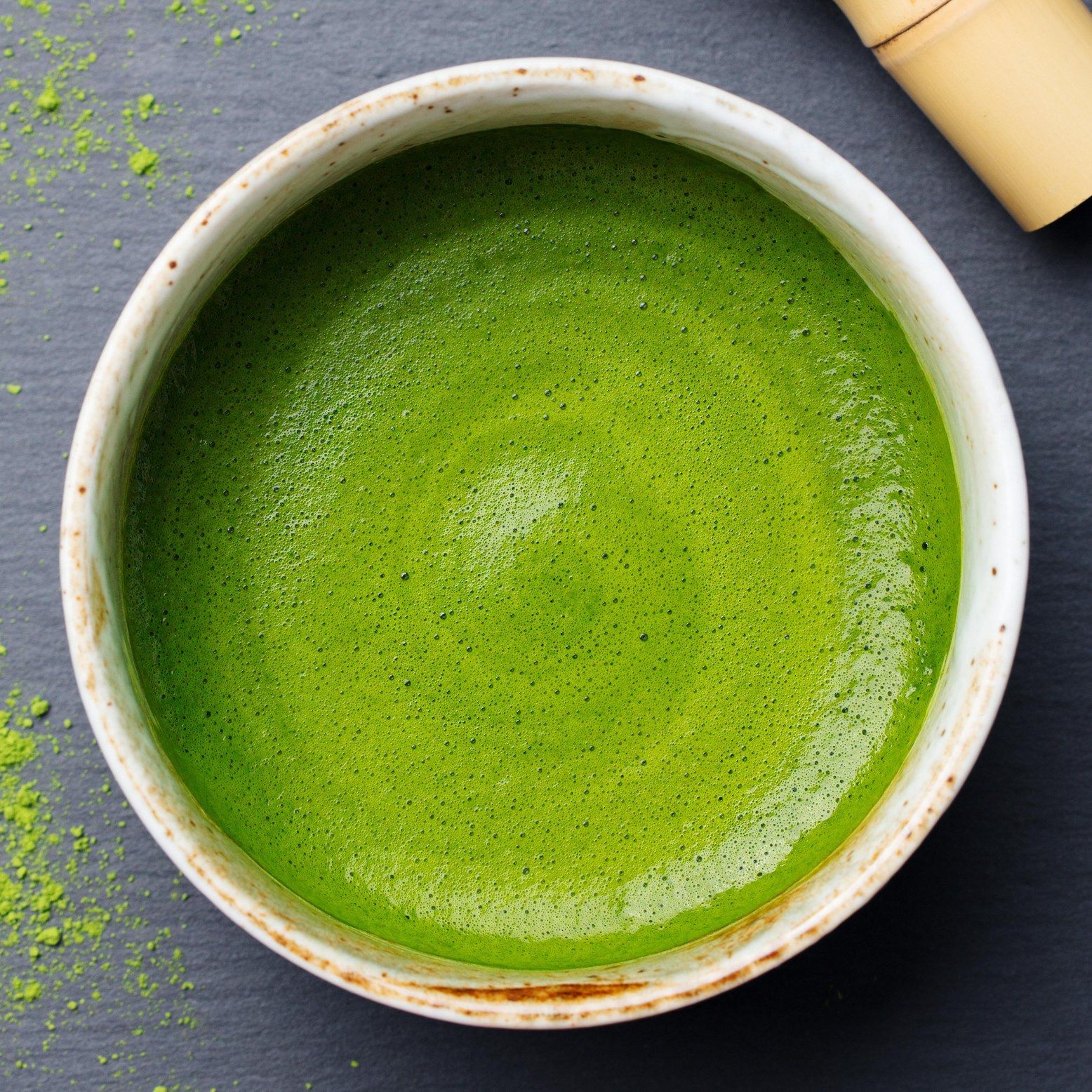 Een kom met opgeklopte matcha thee. De thee is lichtgroen en schuimig. Er ligt een bamboe klopper naast.