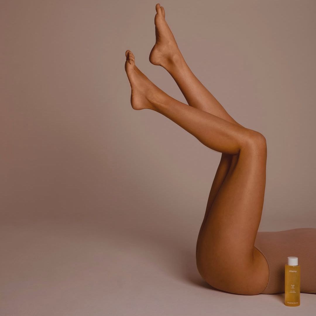 Jambes d'une personne avec un flacon d'huile Alkemy Karité à côté.