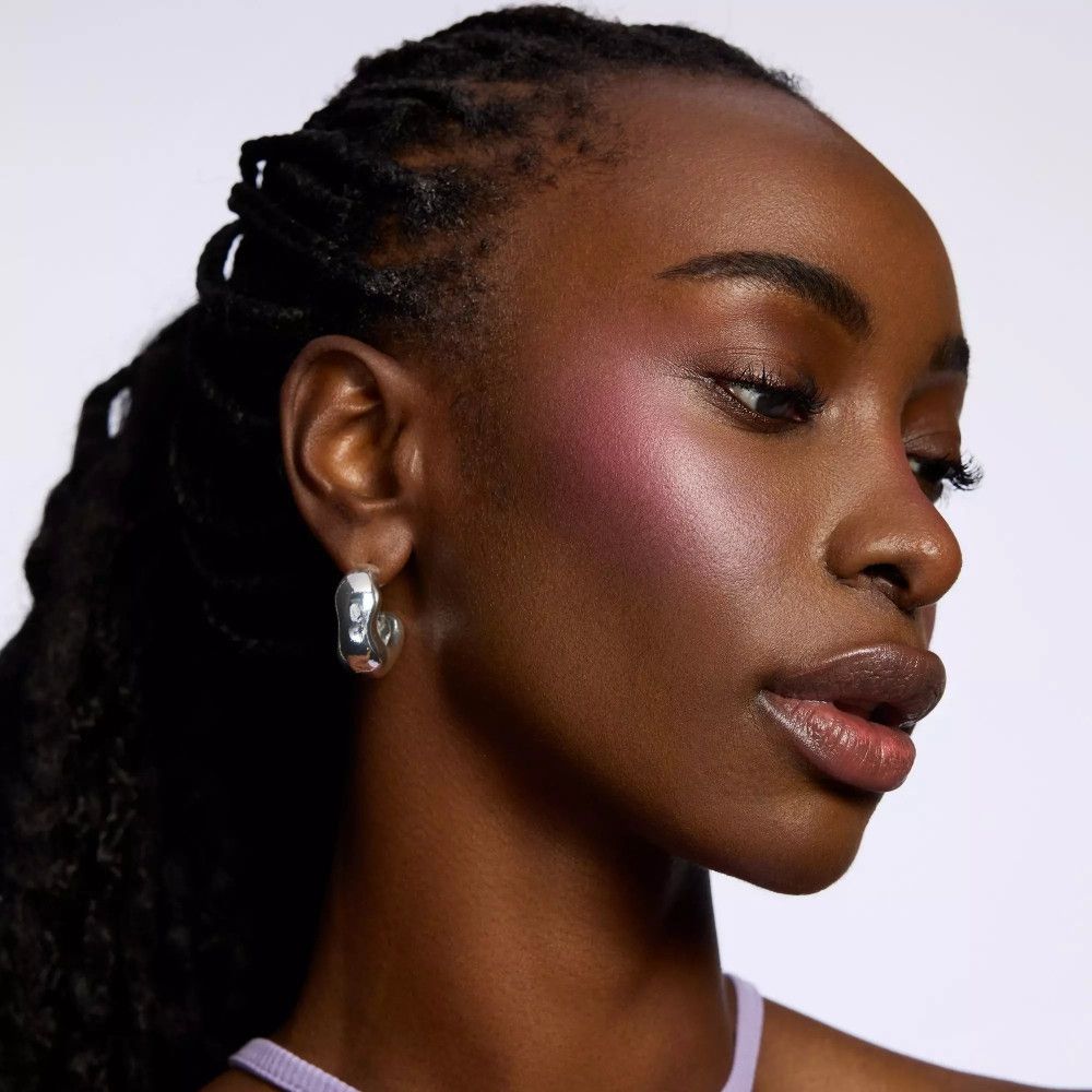 Portrait d'une femme avec du blush rose sur les joues. Elle porte un haut violet et a des tresses. Créoles argentées.