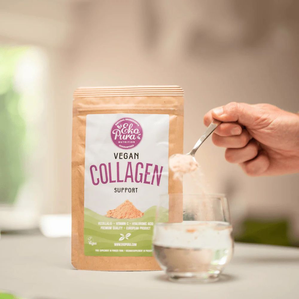Zak met opschrift Vegan Collagen Support. Lepel met poeder boven glas. Hand houdt lepel vast. Achtergrond: keuken.