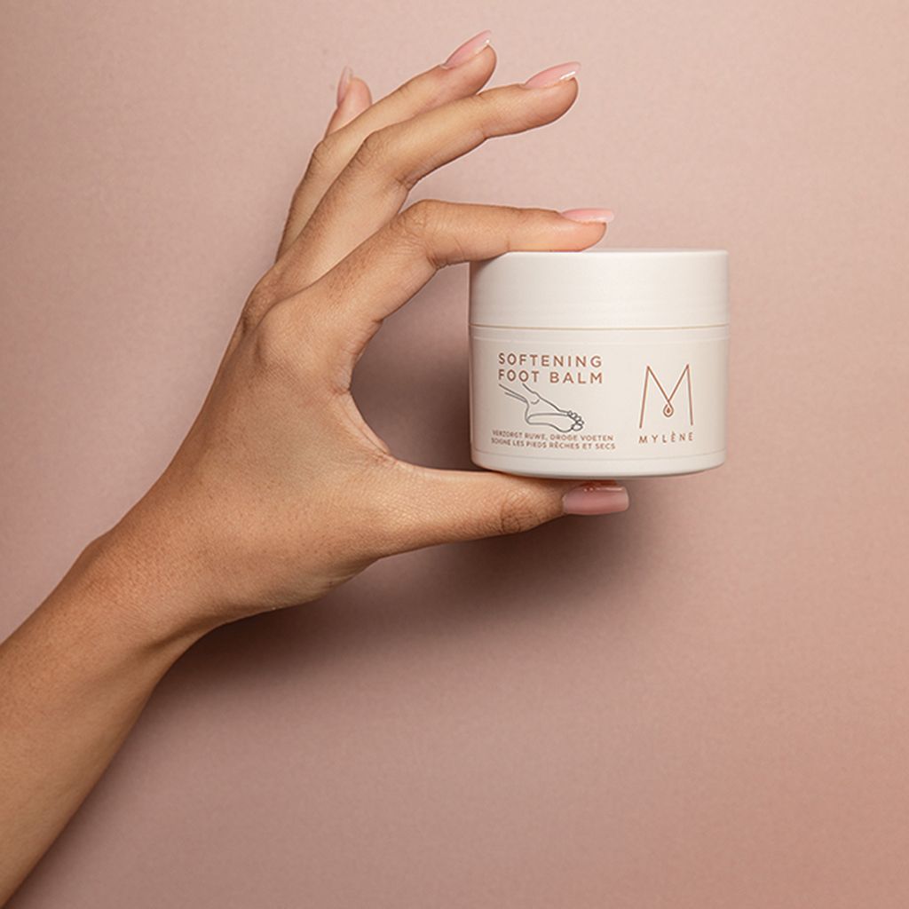 Main tenant un pot blanc avec texte "SOFTENING FOOT BALM" et logo. Texte en dessous : "VERZORGT RUWE, DROGE VOETEN, SOIGNE LES PIEDS RÊCHES ET SECS".
