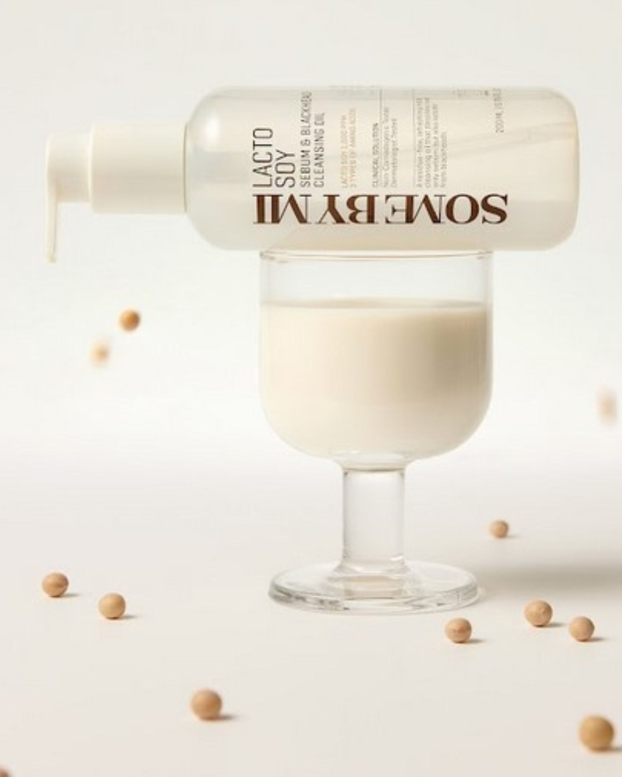 Flacon transparent avec pompe, rempli de liquide, sur un verre. Inscription : SOMEBYMI Lacto Soy Sebum & Blackhead Cleansing Oil. Grains de soja éparpillés.