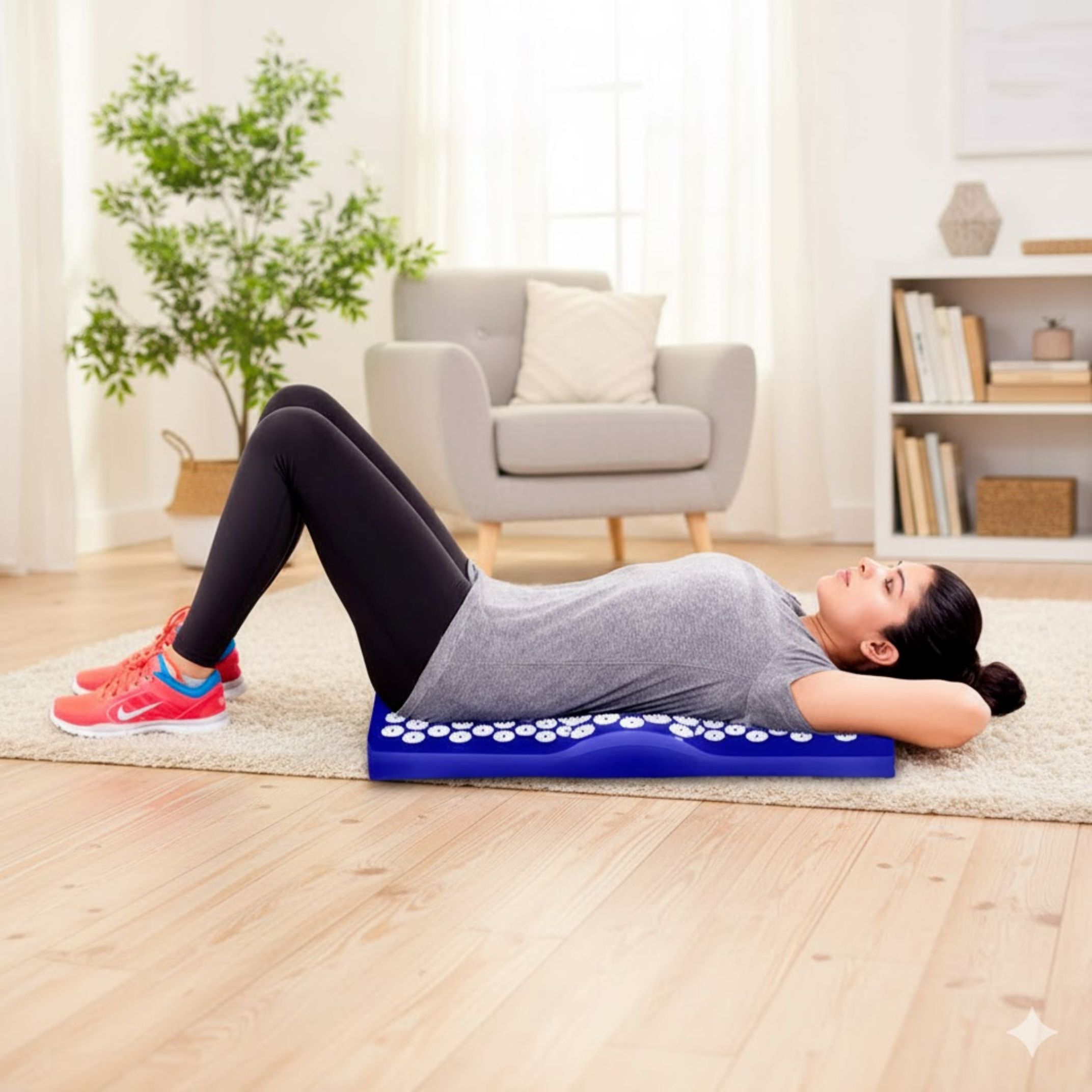 Vrouw ligt op blauwe acupressuurset op tapijt. Mat met witte noppen. Lichte omgeving. Grijs T-shirt.