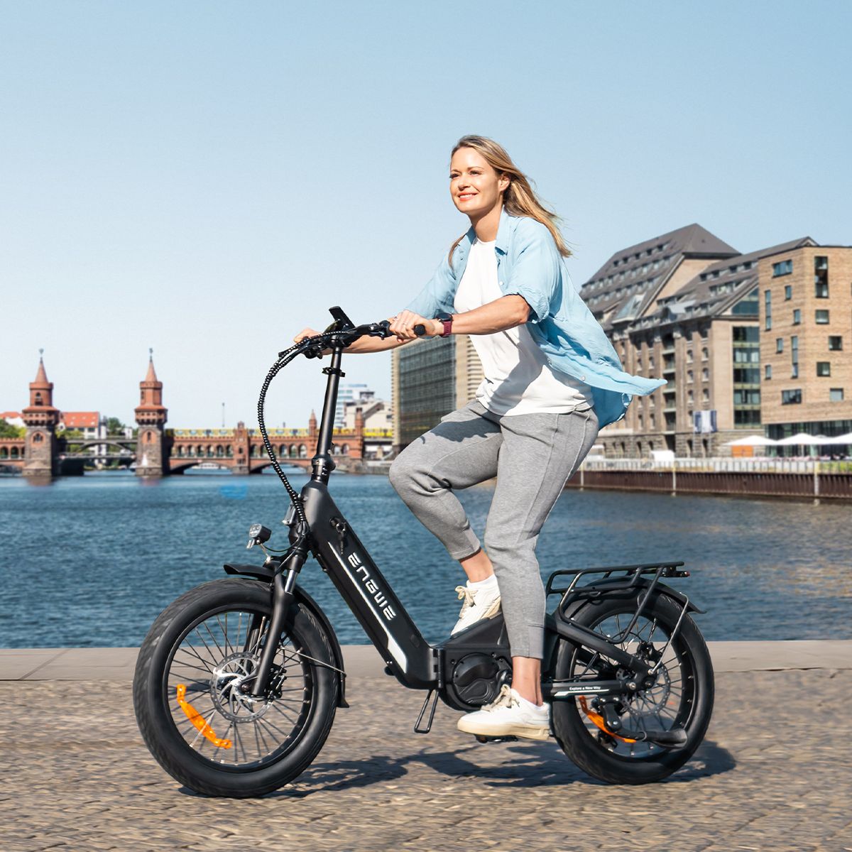 Vrouw die fietst op een zwarte ENGWE L20 3.0 Pro elektrische fiets. Achtergrond: water en gebouwen.