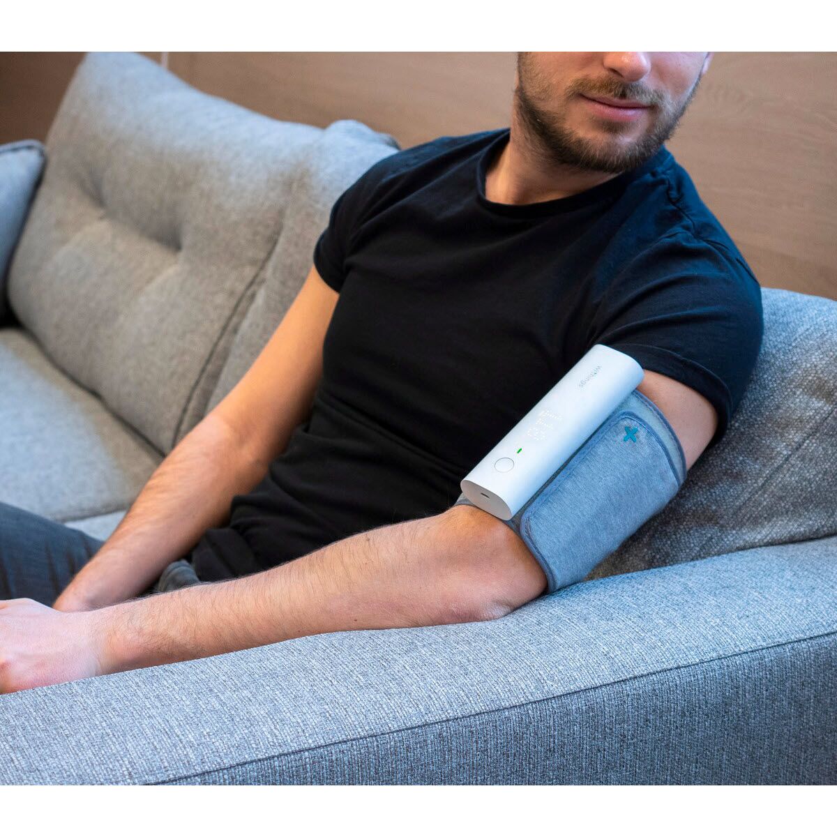 Persoon zit op een bank, arm met grijze bloeddrukmeter. Apparaat met display en Withings-logo.