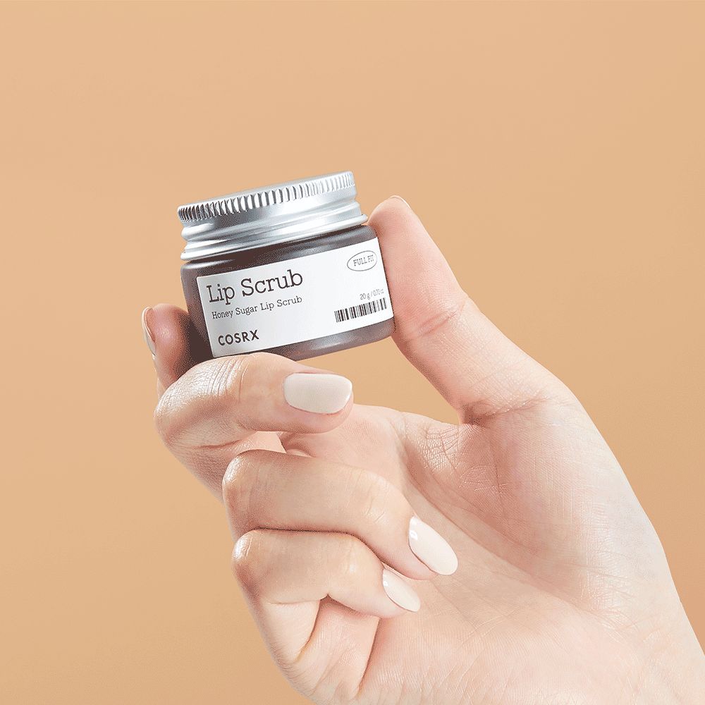 Een hand die een klein potje vasthoudt, gelabeld "Lip Scrub Honey Sugar Lip Scrub COSRX".