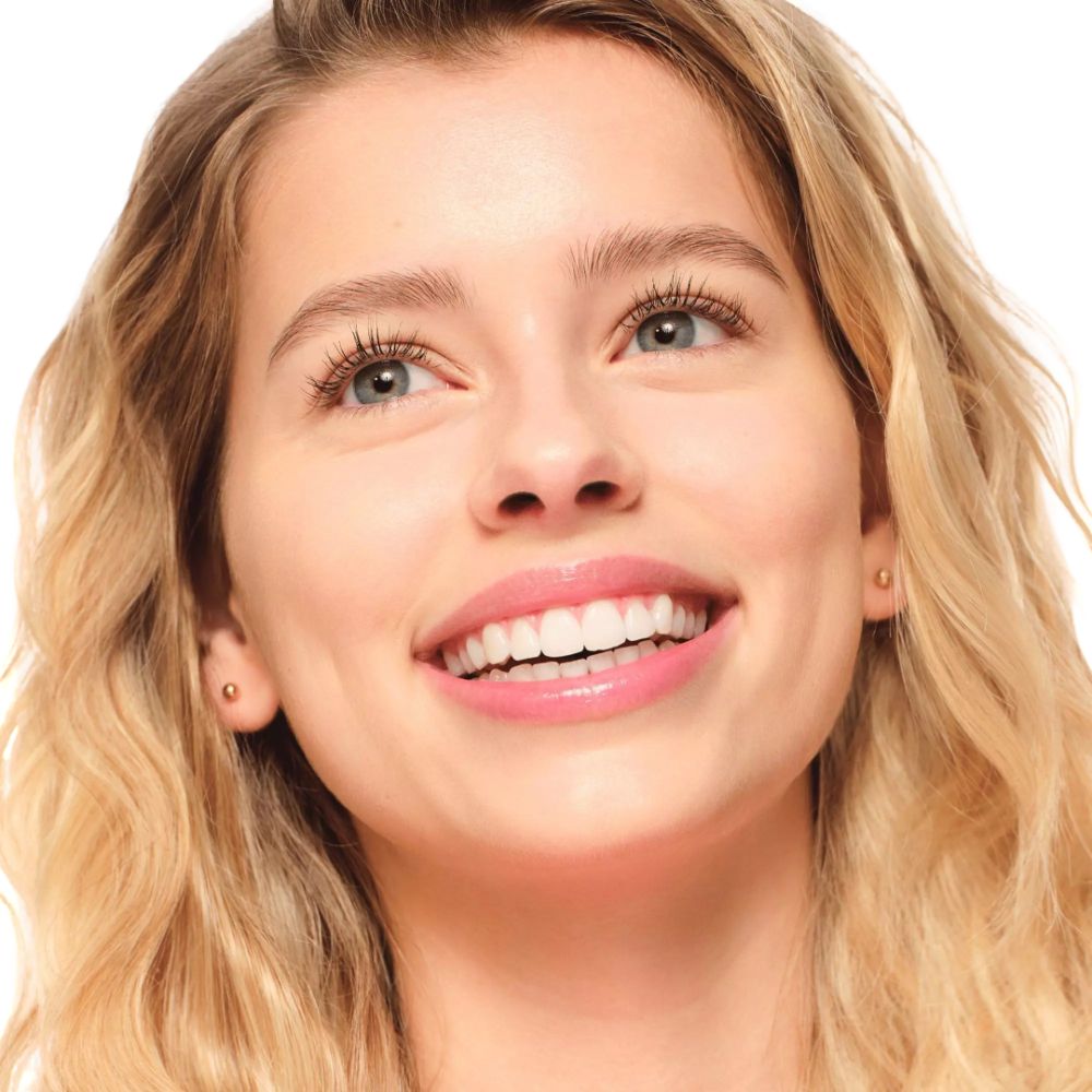 Portrait d'une femme souriante aux cheveux blonds et aux cils maquillés. Elle regarde vers le haut.