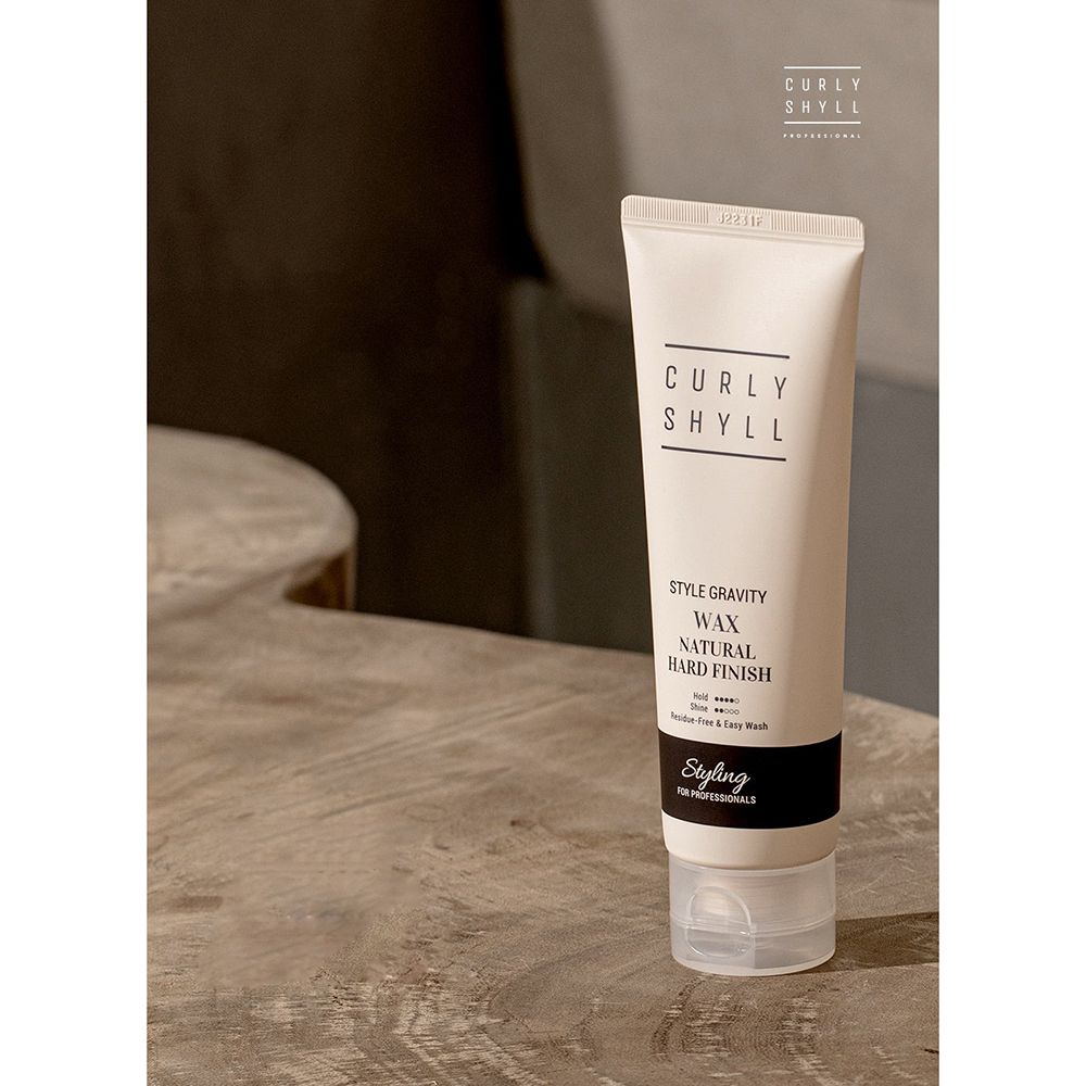 Beige tube met zwarte tekst. Boven: CURLY SHYLL. Daaronder: STYLE GRAVITY WAX NATURAL HARD FINISH. Onderaan: Styling FOR PROFESSIONALS. Op houten oppervlak.