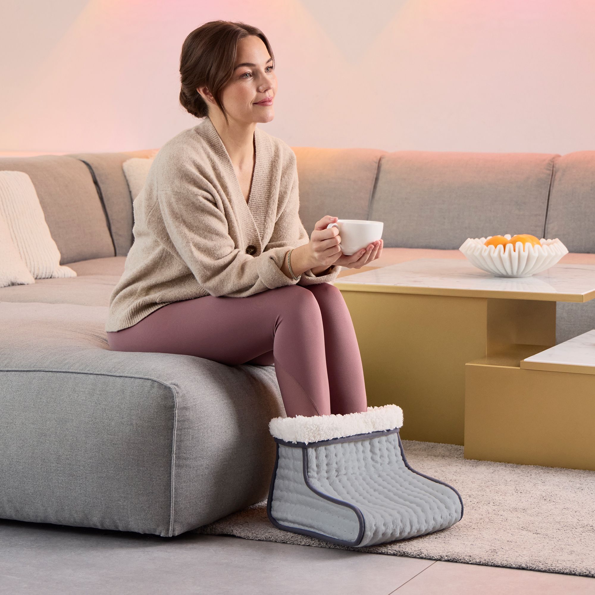 Une femme est assise sur un canapé et tient une tasse. Ses pieds sont dans un chauffe-pieds gris avec une doublure intérieure blanche.