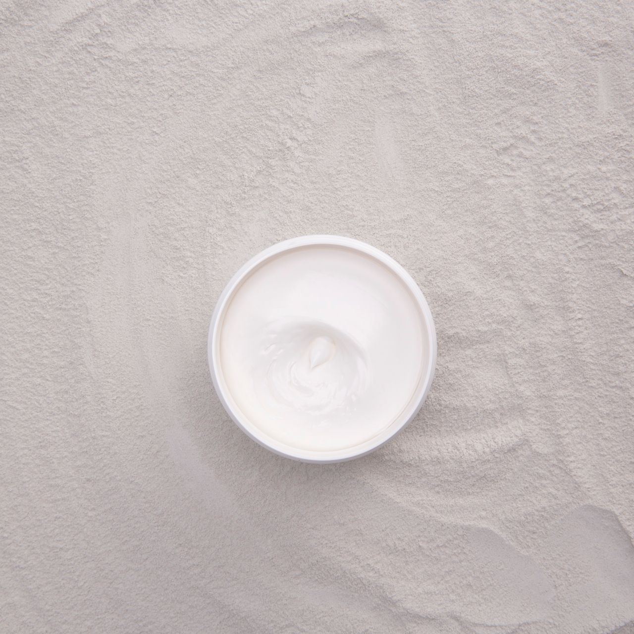 Een witte crème in een open container op een zanderige ondergrond.