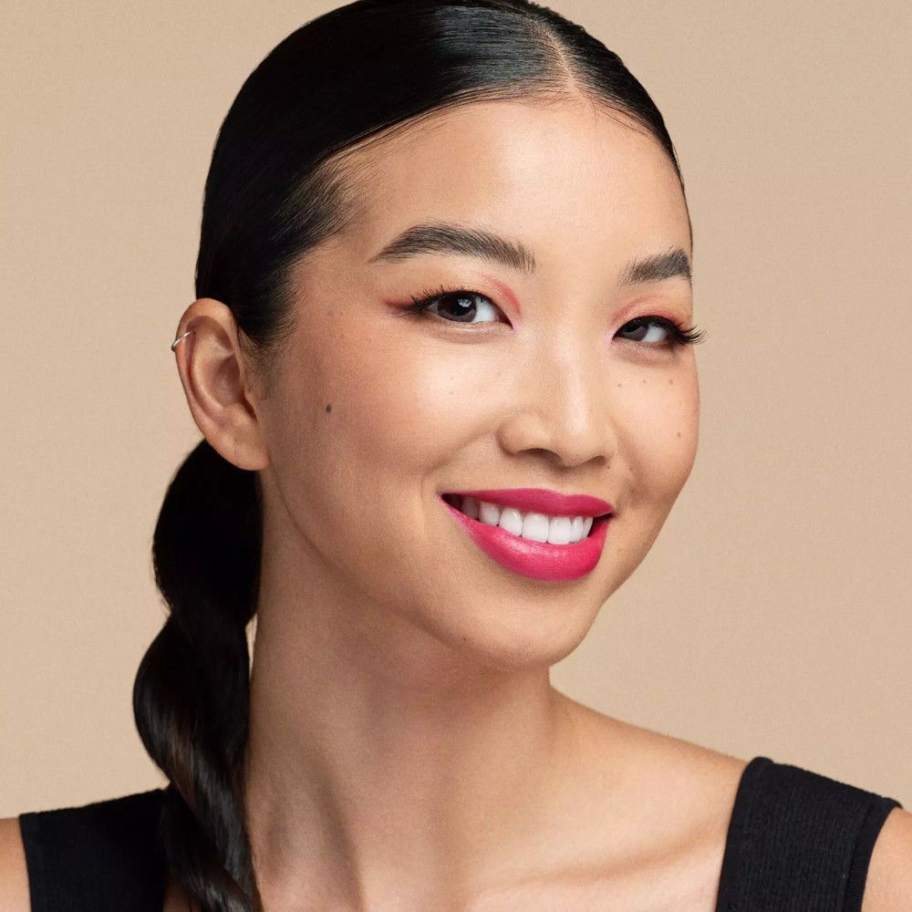 Portret van een lachende vrouw met roze lippenstift en oogschaduw. Ze draagt een zwarte top. Haar haar is zwart.