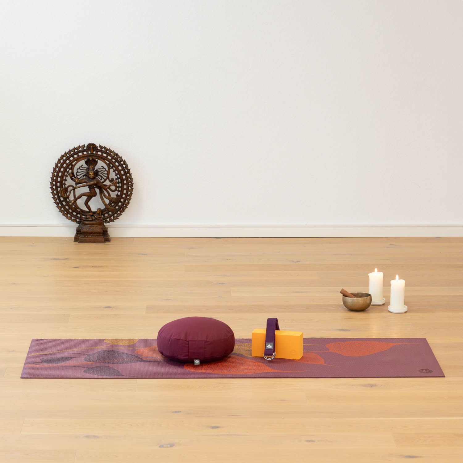 Yogamat met bladerenpatroon, accessoires. Paarse mat met kussen, blok, riem, kaarsen en kom op houten vloer.