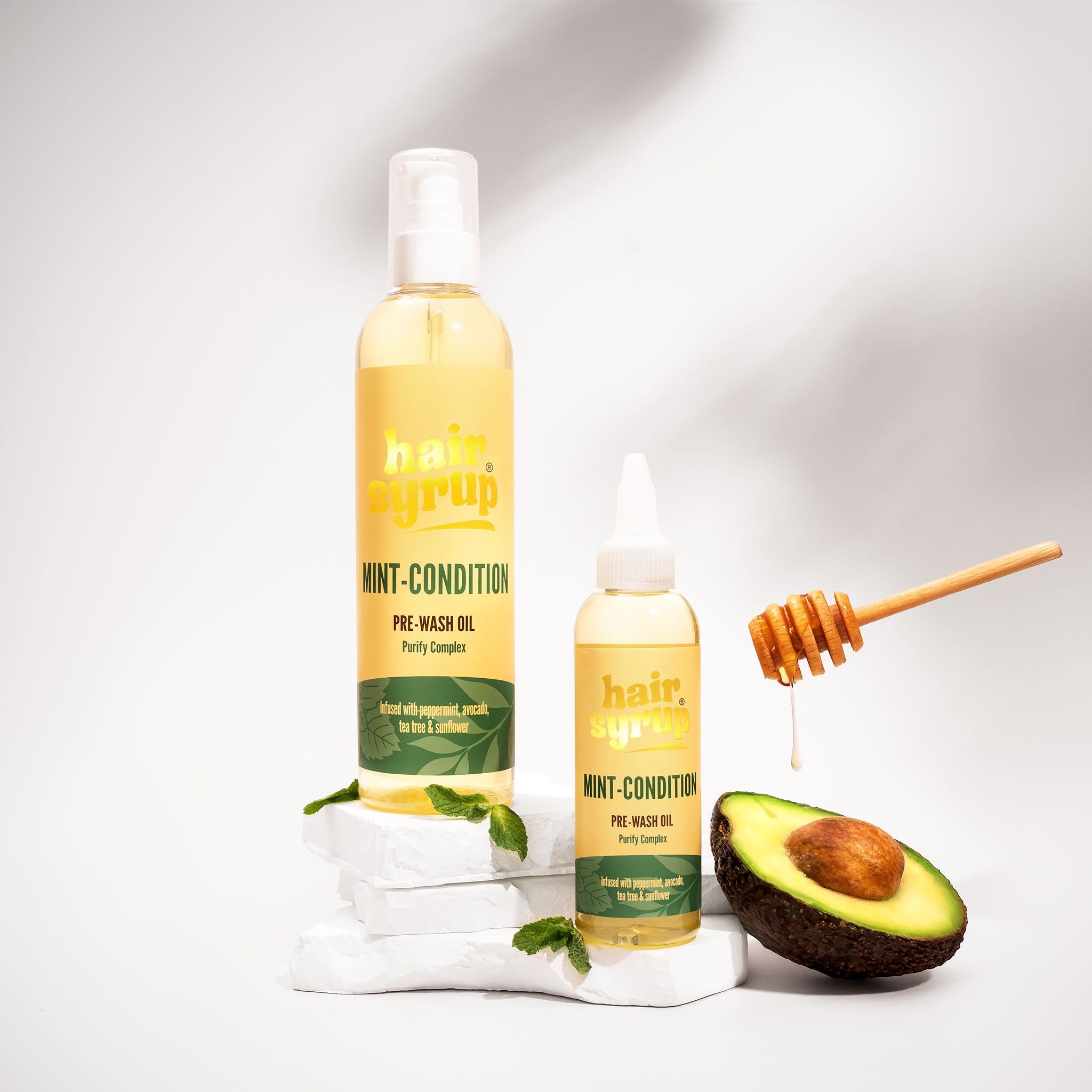 Twee flessen "Hair Syrup Mint Condition" naast avocado en honing. Op witte stenen.