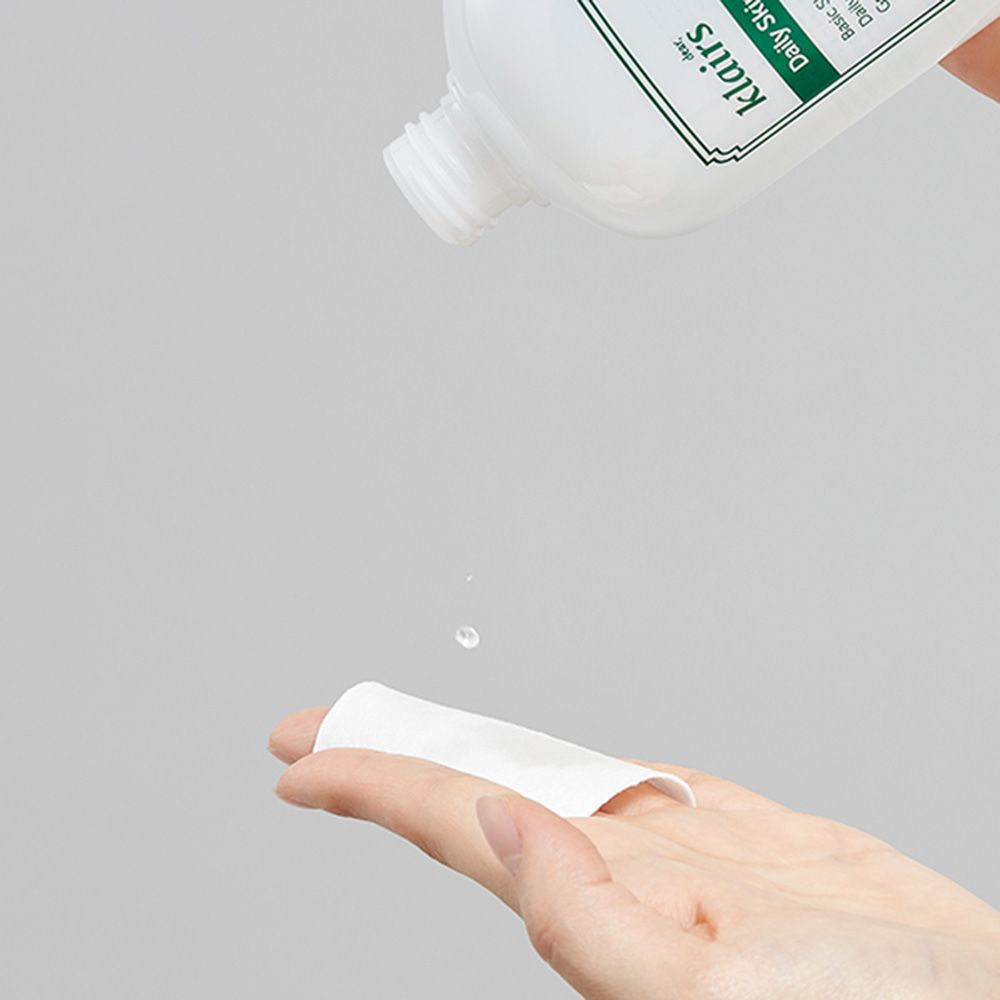 Hand houdt watje vast waar vloeistof uit druppelt. Fles met etiket op de achtergrond. Tekst: klairs, Daily Skin Hydrating Water.