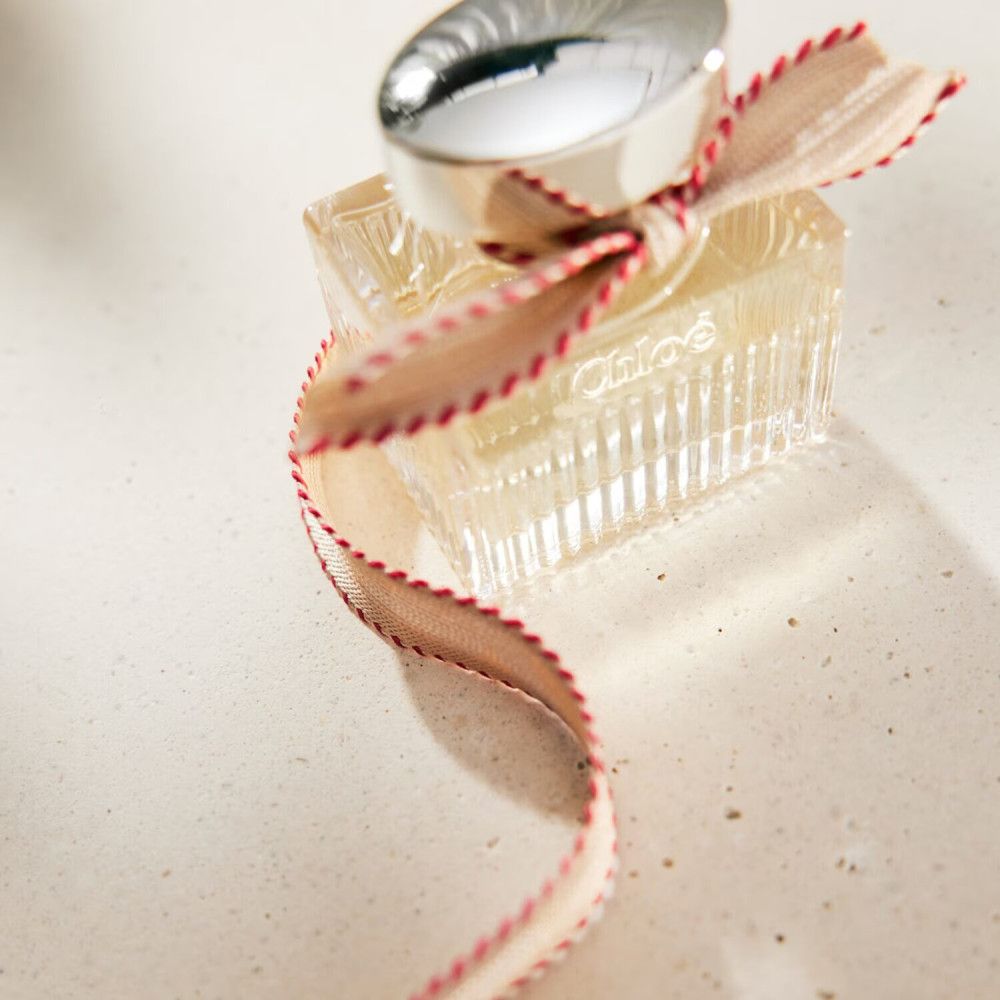 Flacon de parfum avec bouchon argenté et ruban. Le flacon est carré et transparent. Le nom n'est pas visible.