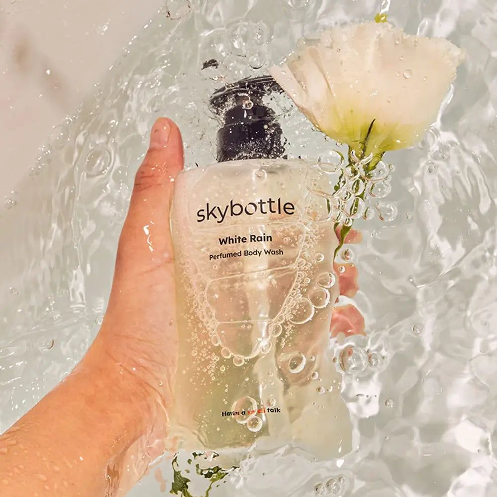 Transparante fles SKYBOTTLE White Rain douchegel met zwarte pomp, in water, met bloem.