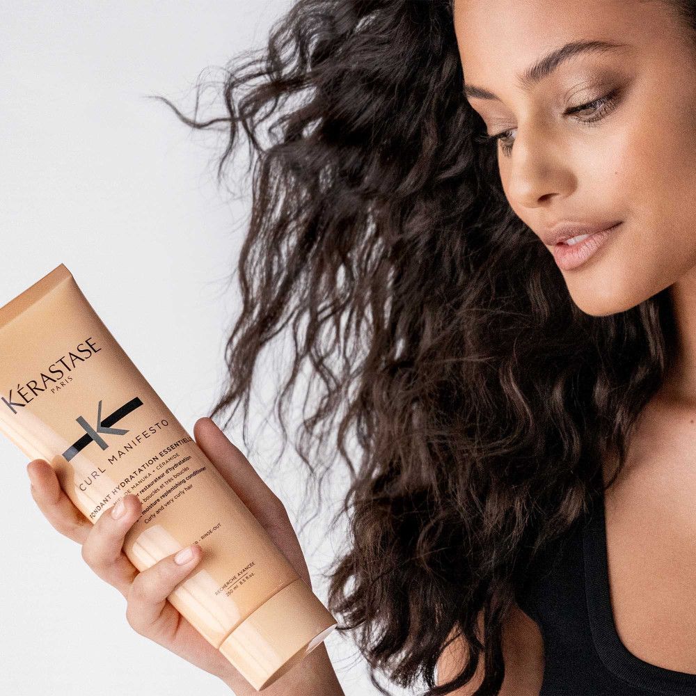 Vrouw houdt een beige tube vast. Kérastase-logo en Curl Manifesto. Productnaam: Fondant Hydratation Essentielle. Krullend haar.