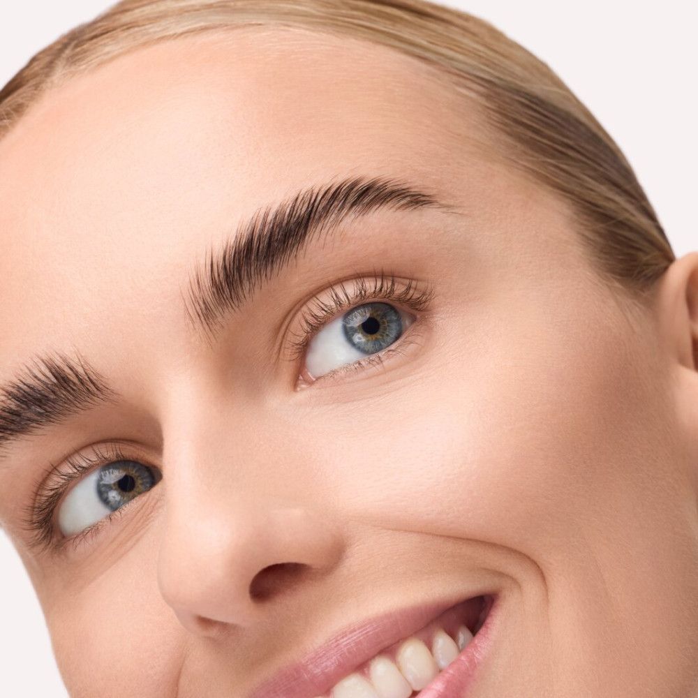 Close-up van een gezicht met geaccentueerde wenkbrauwen. De wenkbrauwen zijn naar boven geborsteld en lijken voller. Blauwe ogen, lichte huid.