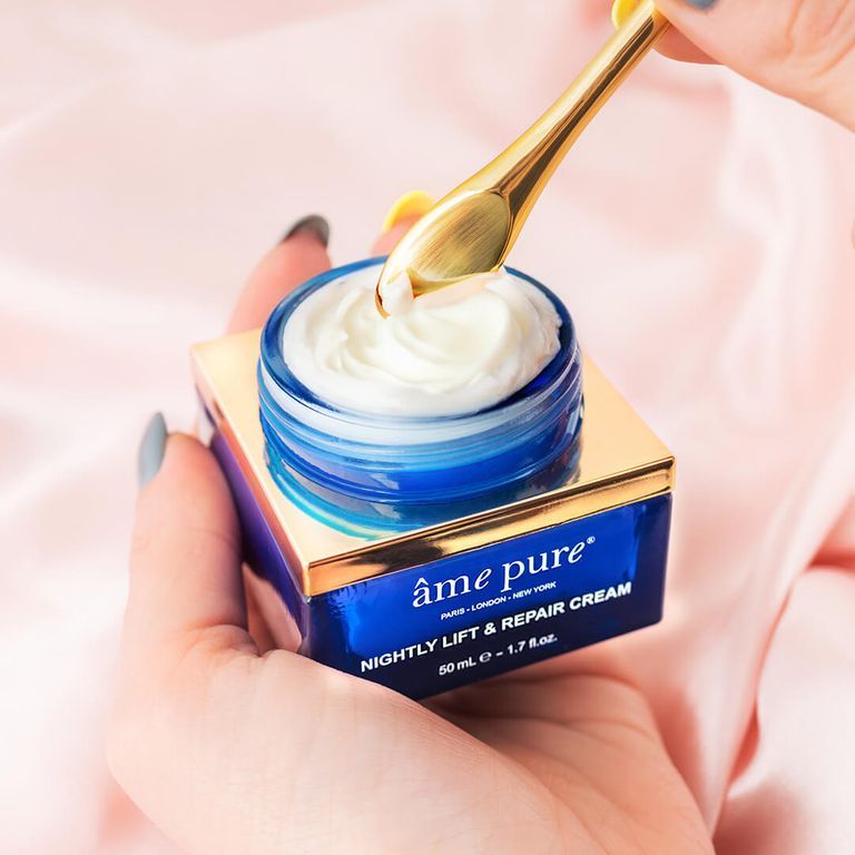 Een hand schept crème uit een potje âme pure Nightly Lift & Repair met een gouden spatel.
