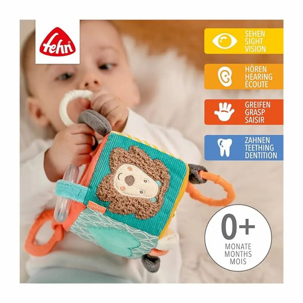 Baby houdt stoffen kubus met leeuwenmotief vast. Merk 'Fehn' zichtbaar. Indicaties voor zien, horen, grijpen, tanden. Leeftijd: 0+ maanden.