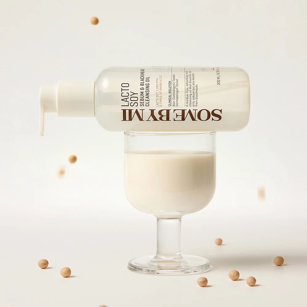 Flacon transparent avec pompe, rempli de liquide, sur un verre. Inscription : SOMEBYMI Lacto Soy Sebum & Blackhead Cleansing Oil. Grains de soja éparpillés.