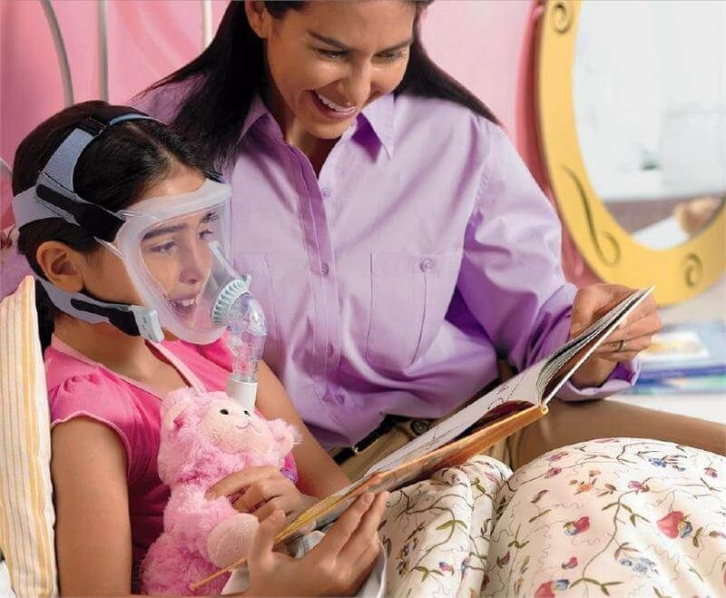 Enfant portant un masque facial complet Philips CPAP, mère lisant, dans une chambre.