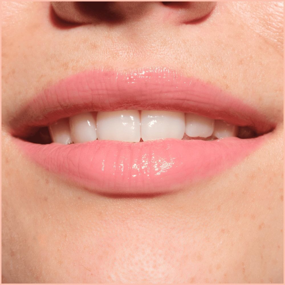 Gros plan sur les lèvres avec repulpeur appliqué. Teinte rose, fini brillant. Dents clairement visibles.