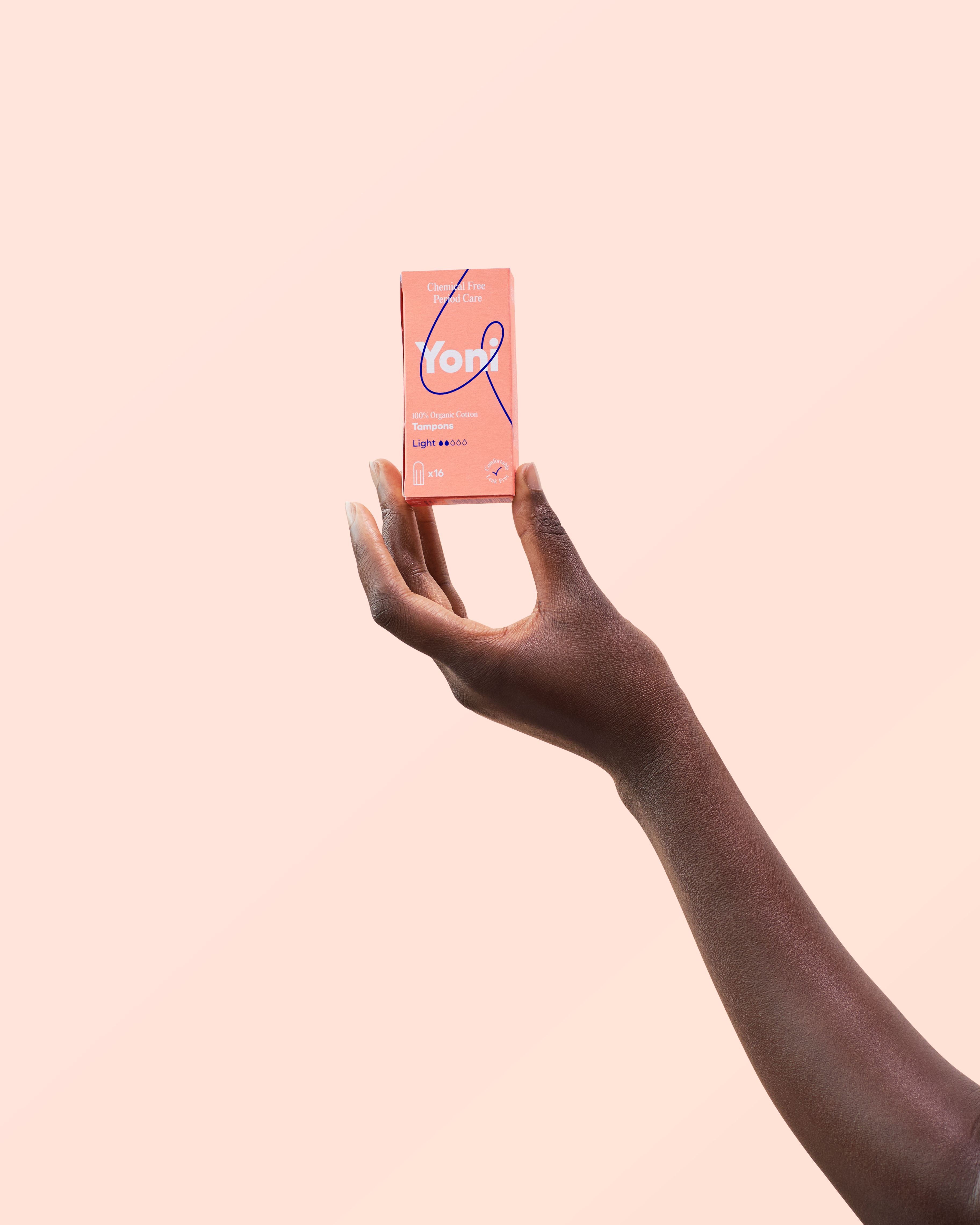 Hand houdt een pakje tampons vast. Opschrift: Yoni, 100% biologisch katoen, Light. Achtergrond: lichtroze.