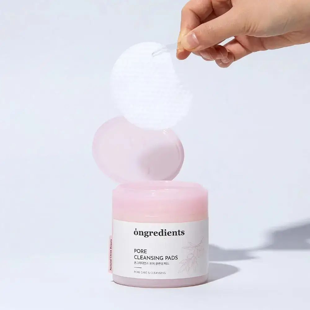 Ronde pot met roze deksel. Opschrift: Ongredients, Pore Cleansing Pads. Hand houdt pad vast.