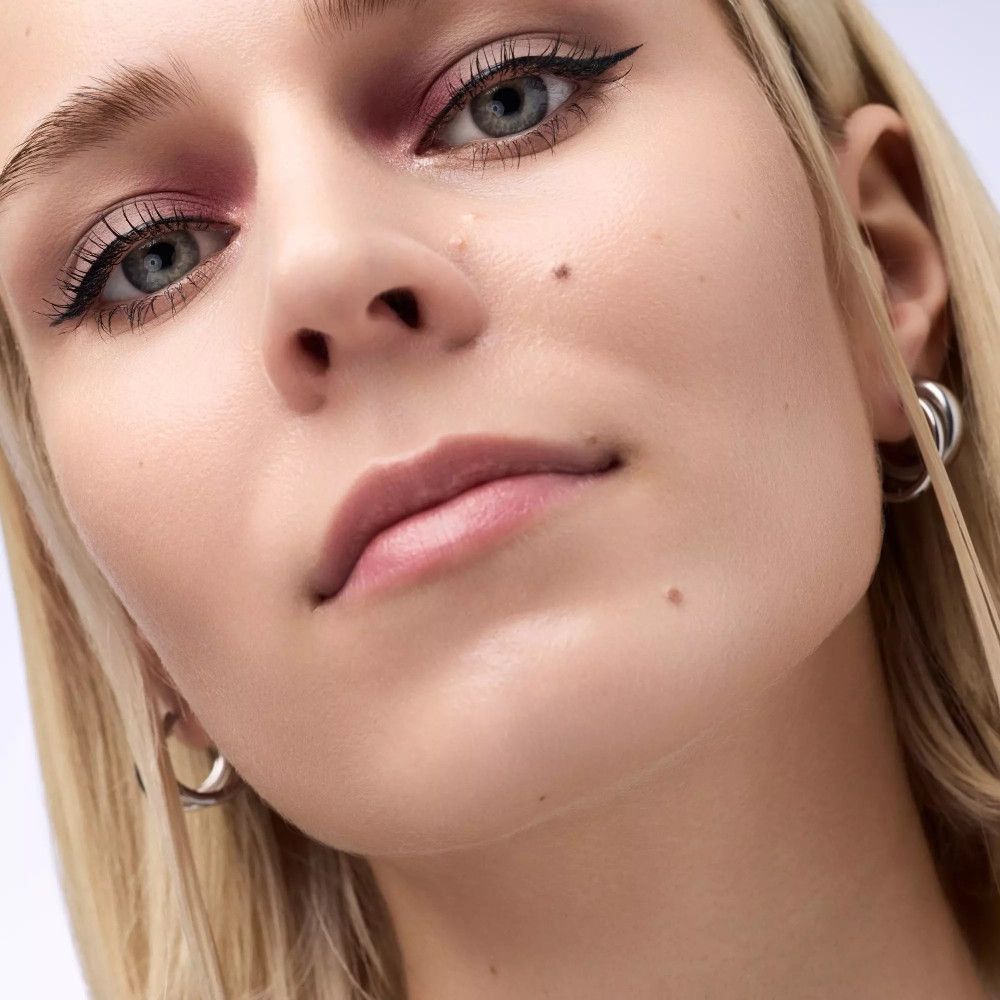 Gros plan sur un visage avec fard à paupières rose et eye-liner noir. Cheveux blonds et boucles d'oreilles argentées.