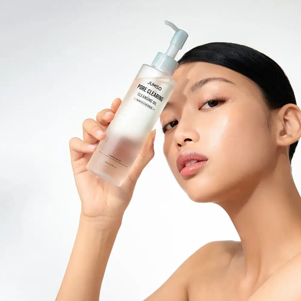 Vrouw houdt een transparante fles met pomp vast. Opschrift: JUMISO PORE CLEARING CLEANSING OIL. Serum Control.