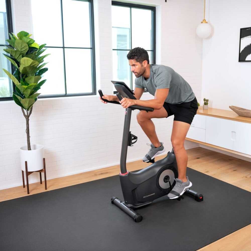 Homme sur un vélo d'exercice noir. Logo Schwinn. Dans un salon. Tapis noir. Vêtements gris.