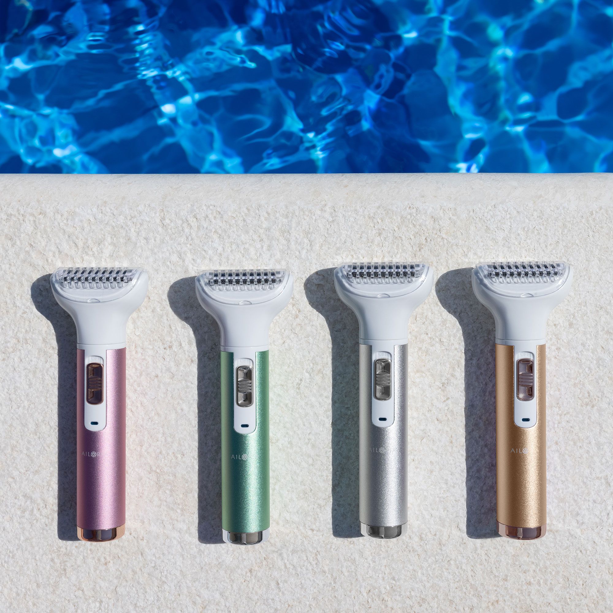 Vier scheerapparaten met gekleurde handvatten. Roze, groen, zilver en goud. Liggend op een wit oppervlak bij water.