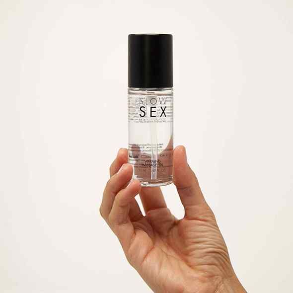 Main tenant un flacon d'huile de massage. Inscription : SLOW SEX. Intitulé : Warming Massage Oil. Bouchon noir, liquide transparent.
