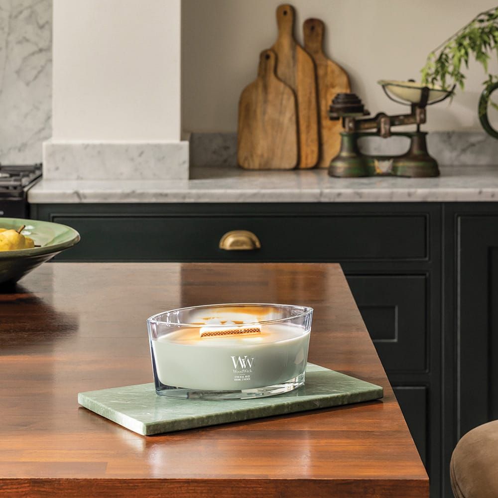 Kaars met houten lont op groene plaat. Glazen houder met groene was. 'WW' logo. Op houten tafel.