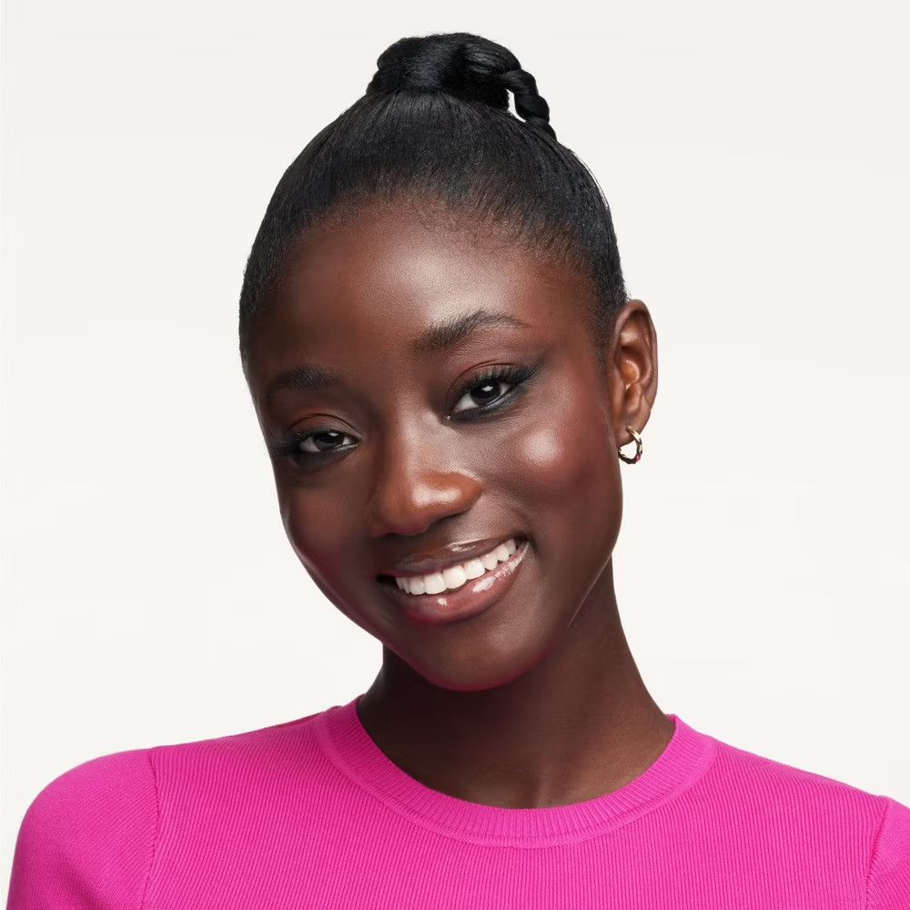 Portrait d'une femme à la peau foncée, cheveux attachés. Elle porte un pull rose et sourit. Elle porte des boucles d'oreilles.