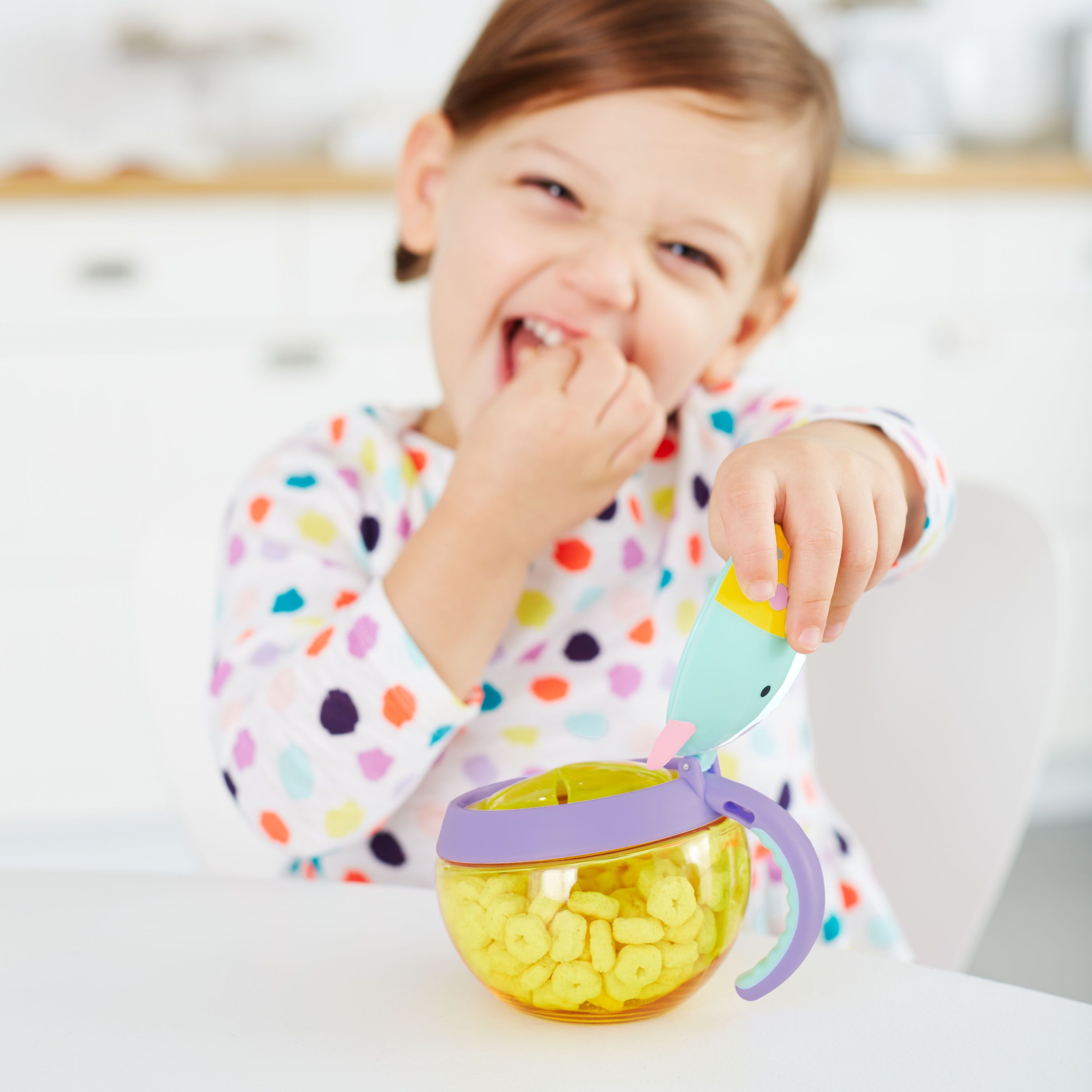 Kind eet, houdt snackbox met eenhoorn design vast. Gele container met snacks, paarse ring, blauw-geel deksel. Met handvat.