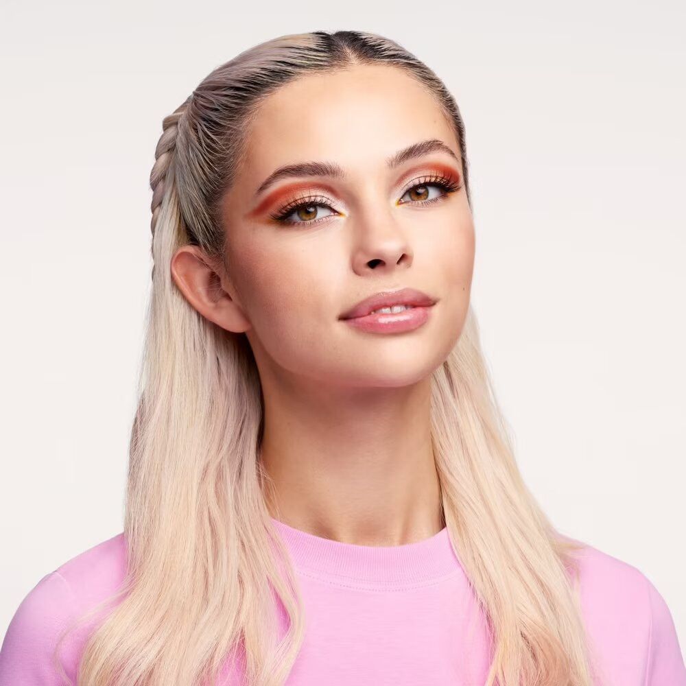 Portrait d'une femme avec des tresses blondes et un haut rose. Elle porte du maquillage. Elle regarde la caméra.