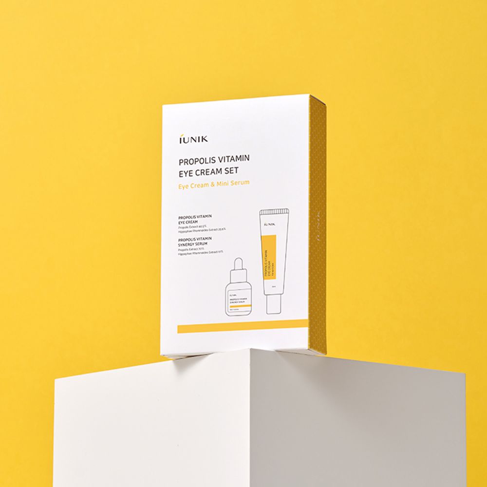 iUNIK Propolis Vitamin Eye Cream Set. Productverpakking op wit voetstuk, gele achtergrond. Tekst op de verpakking.
