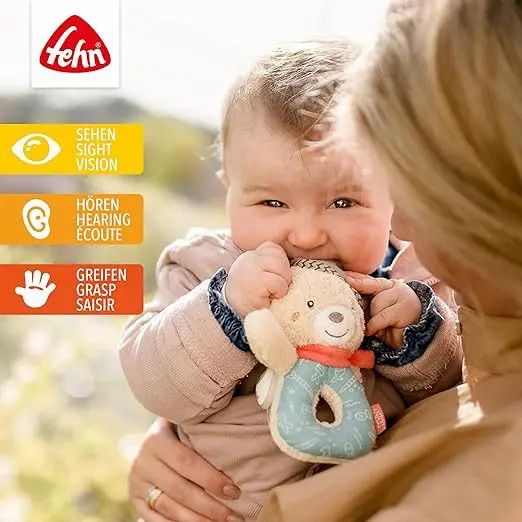 Baby houdt pluche beer rammelaar in de mond. Fehn-logo linksboven. Visuele, auditieve en tactiele iconen.