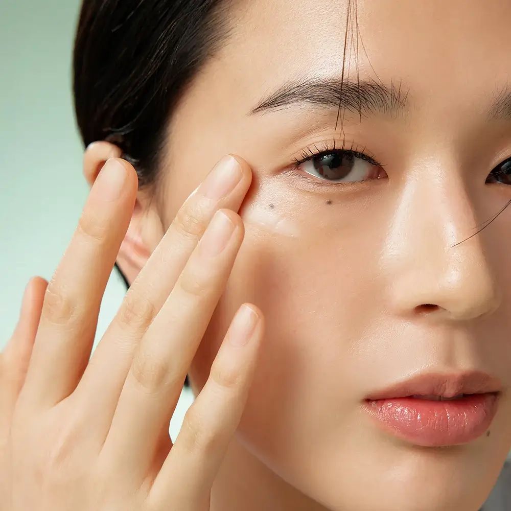 Gros plan d'une personne appliquant de la crème sous l'œil. La main touche doucement la peau. Peau claire, cheveux foncés.